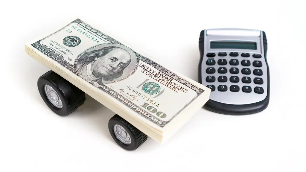 A stack of dollar bills on miniature car wheels beside a calculator.